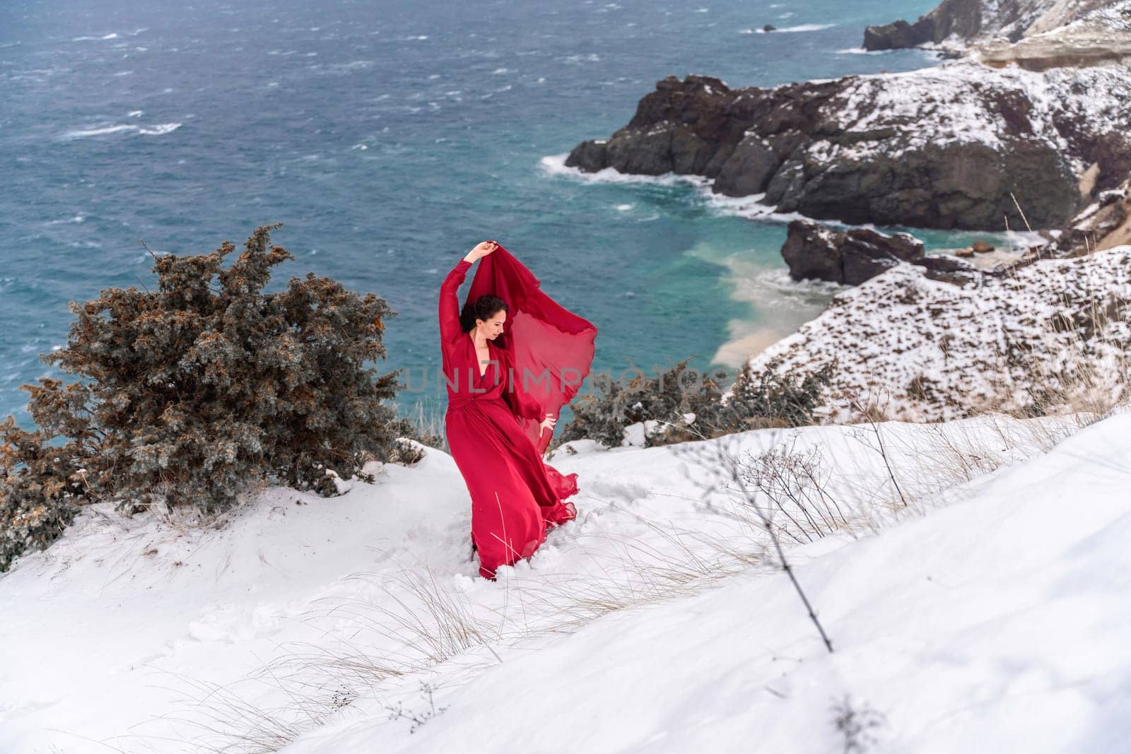 Woman red dress snow sea. Happy woman in a red dress in the snowy mountains by the emerald sea. The wind blows her clothes, posing against sea and snow background. by Matiunina