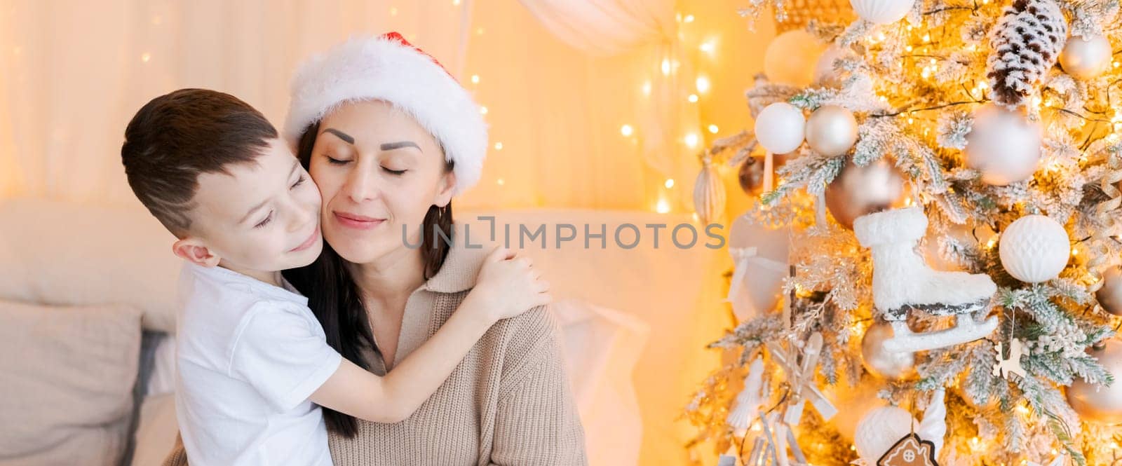 Happy mom and son wearing santa claus hat cuddle on bed in decorated bedroom on Christmas Eve in anticipation of the party