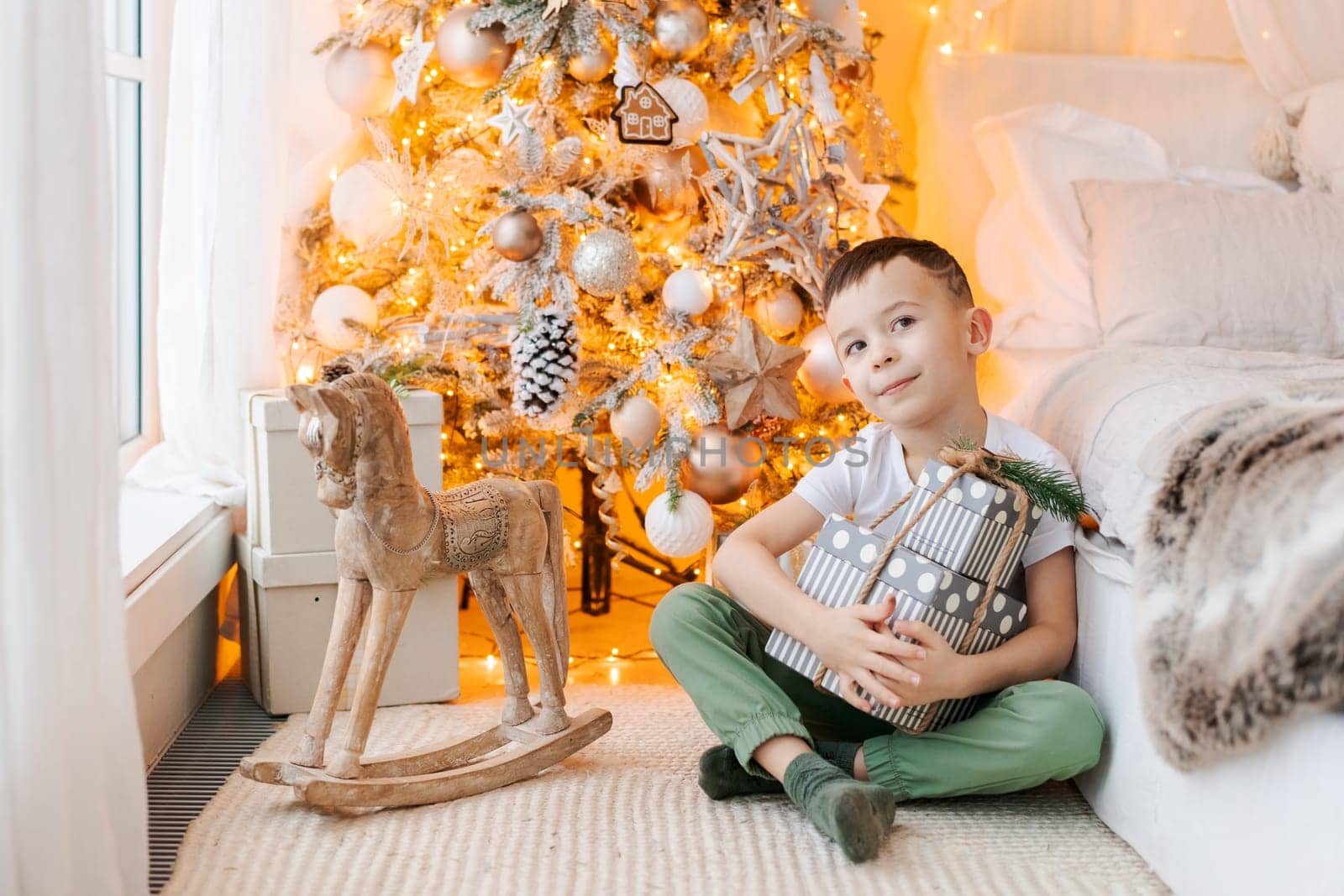 Happy boy sits on floor with gift in his hand by window in front Christmas tree in his bedroom