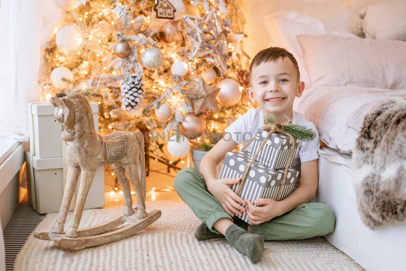 Happy boy sits on floor with gift in his hand by window in front Christmas tree by EkaterinaPereslavtseva