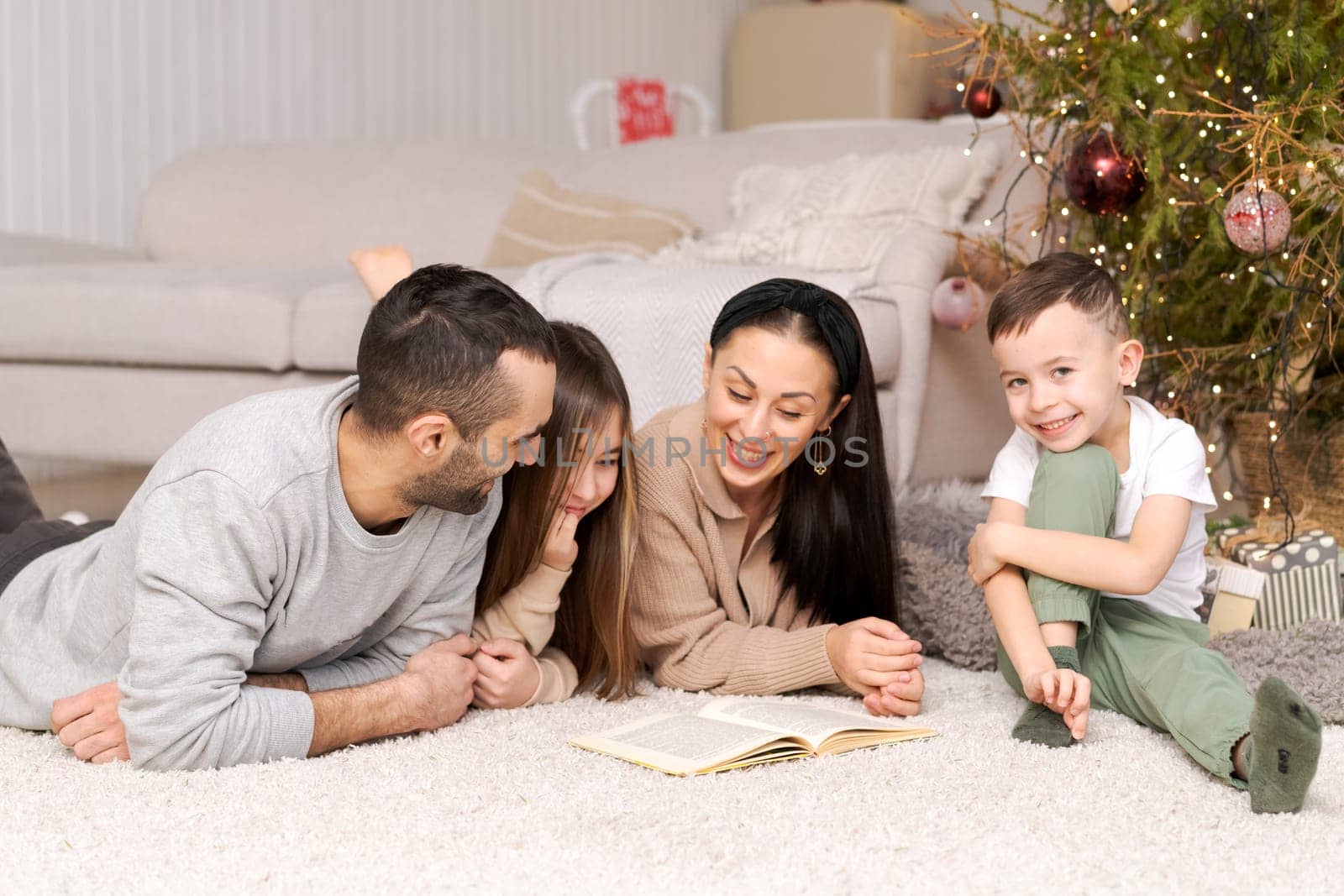 Happy family in decorated house at christmas lying on floor reading book against background of couch and Christmas tree. Selective focus