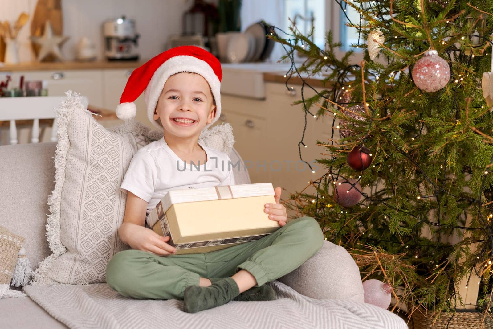 Cute little boy in Santa hat sits on couch with Christmas present from his by EkaterinaPereslavtseva