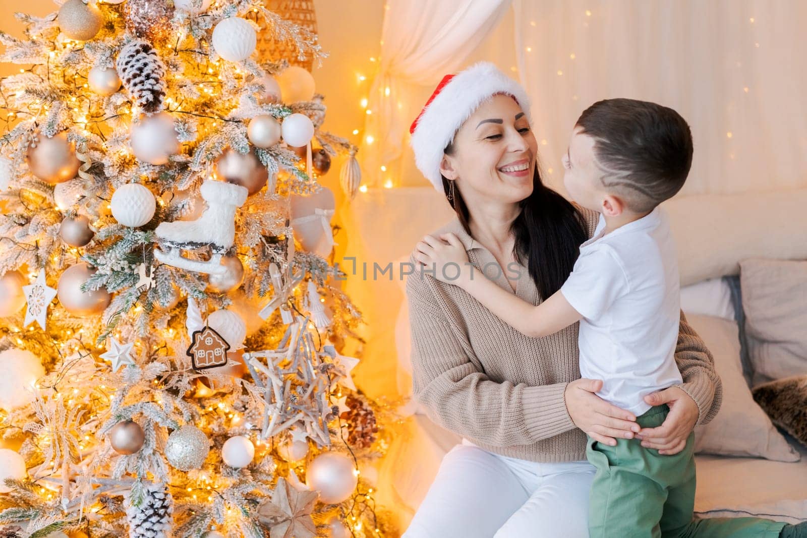 Happy mom and son wearing santa claus hat cuddle on bed in decorated bedroom by EkaterinaPereslavtseva