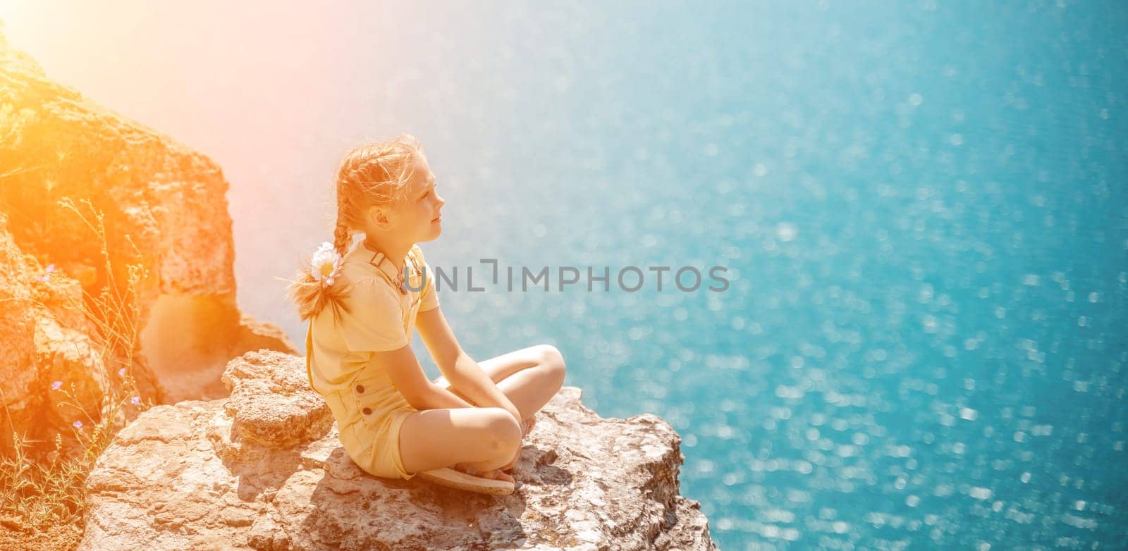 Happy girl perched atop a high rock above the sea, wearing a yellow jumpsuit and braided hair, signifying the concept of summer vacation at the beach. by Matiunina