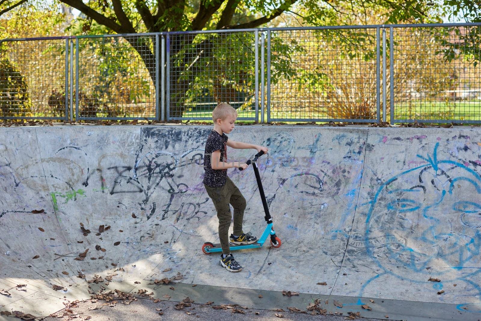 Boy on scooter makes a trick and enjoying his riding in the skate park at cloudy spring day. Young man doing trick on the kick scooter in the park. Full body little boy on the kick scooter on sunny autumn day in skate park. From below of active teen boy with kick scooter standing on ramp in skate park while preparing for performing trick in sunny spring day. by Andrii_Ko