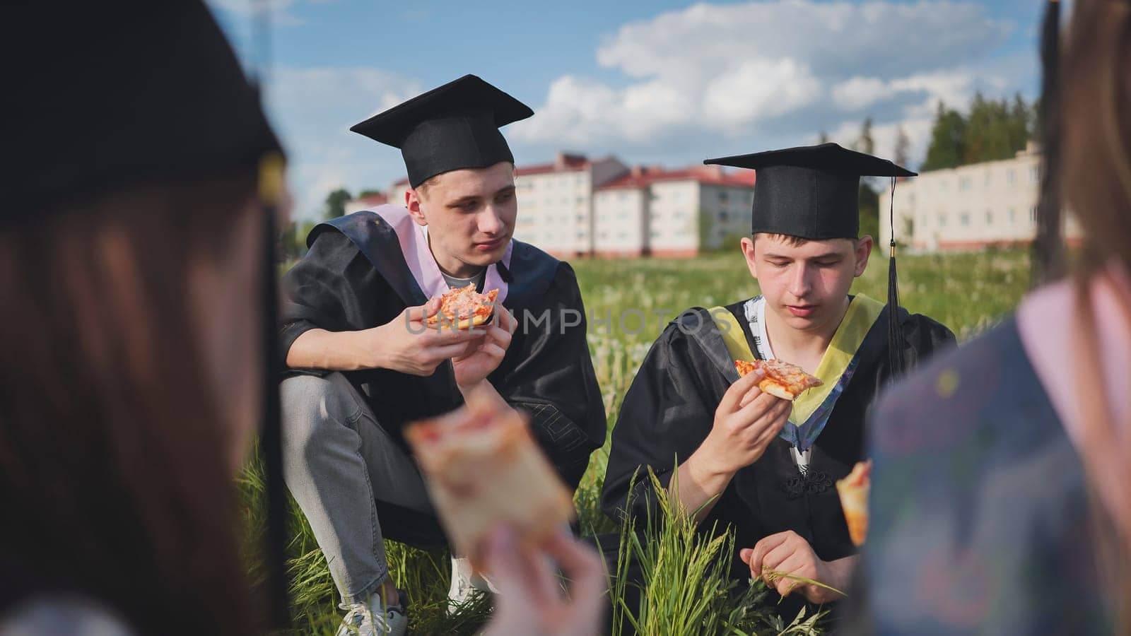 Graduates in black suits eating pizza in a city meadow. by DovidPro