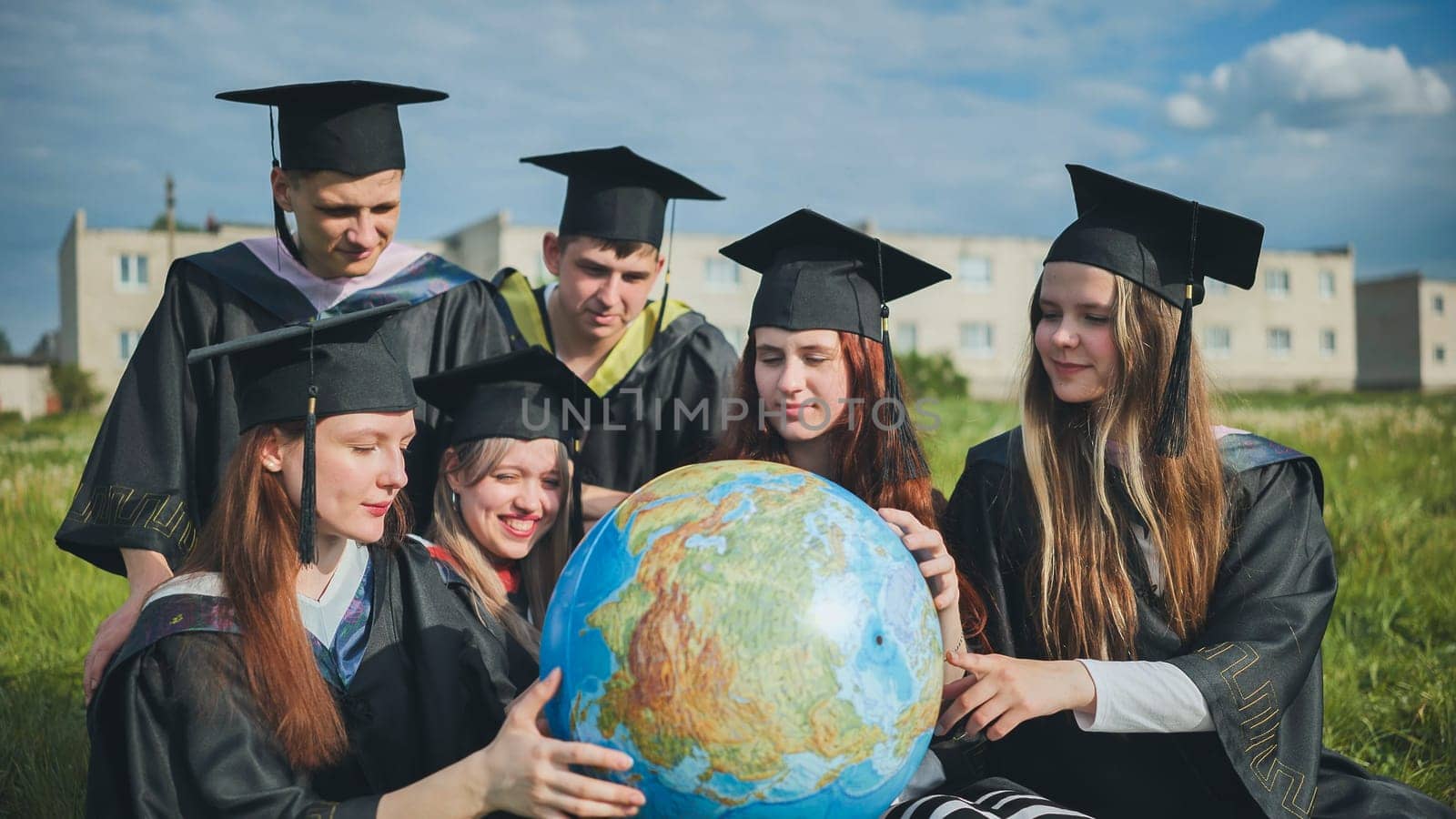 Graduates in black robes examine a geographical globe sitting on the grass. by DovidPro