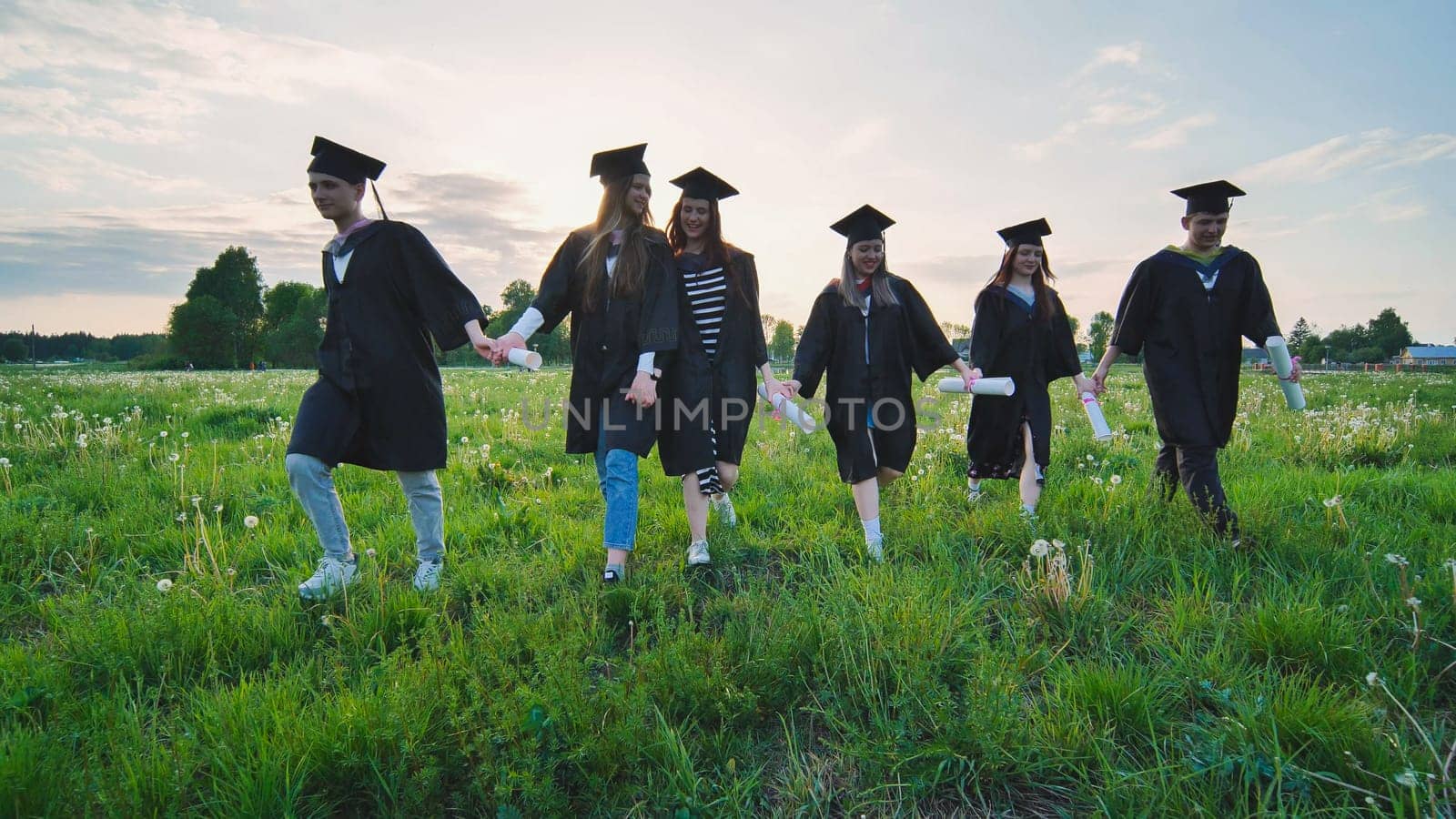 Six graduates in robes walk against the backdrop of the sunset. by DovidPro