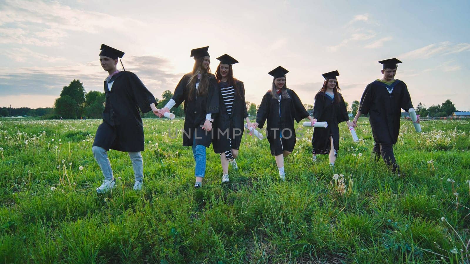 Six graduates in robes walk against the backdrop of the sunset