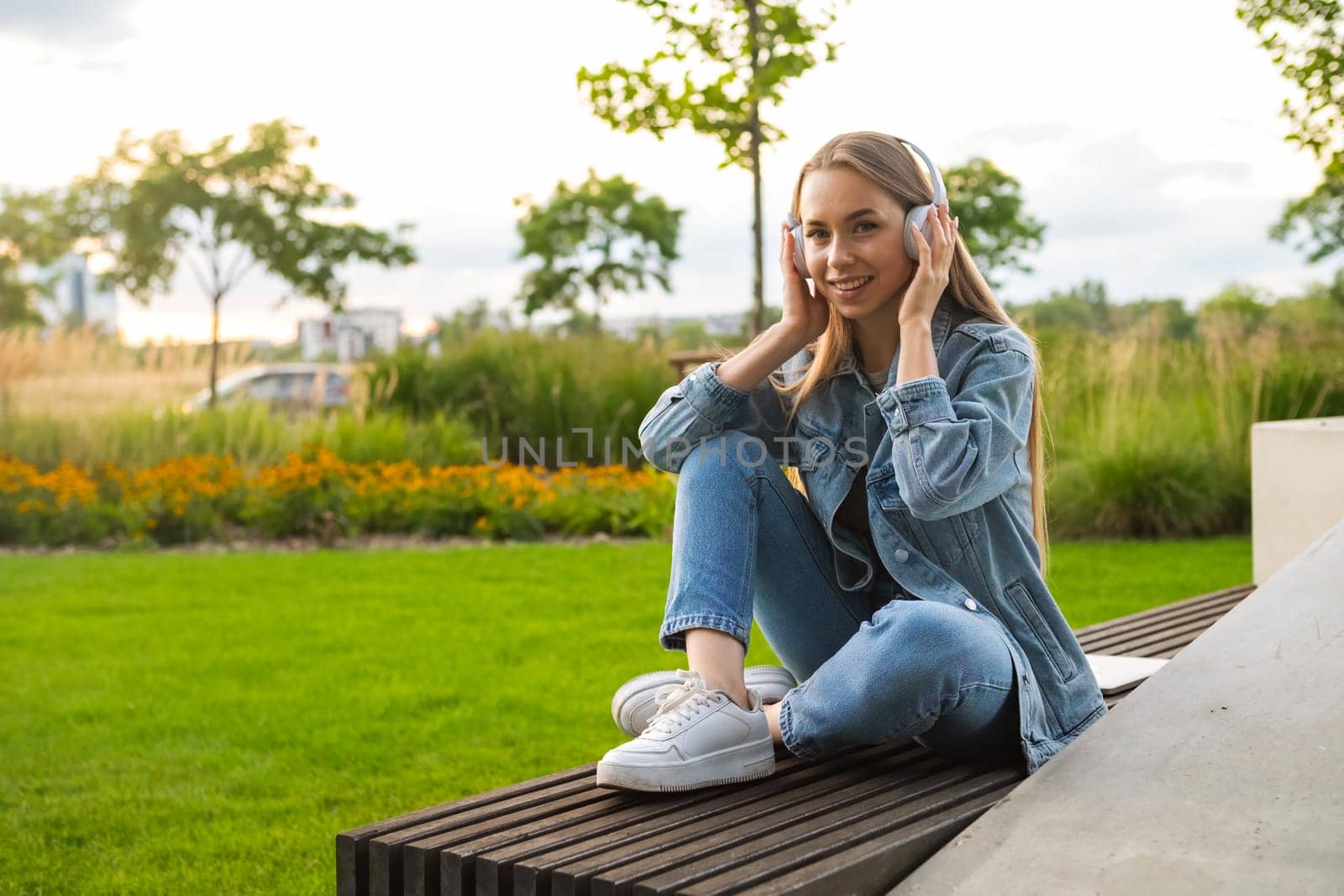 A blonde woman sits on a bench with crossed legs, enjoying through her headphones, enjoying the sunset in the park