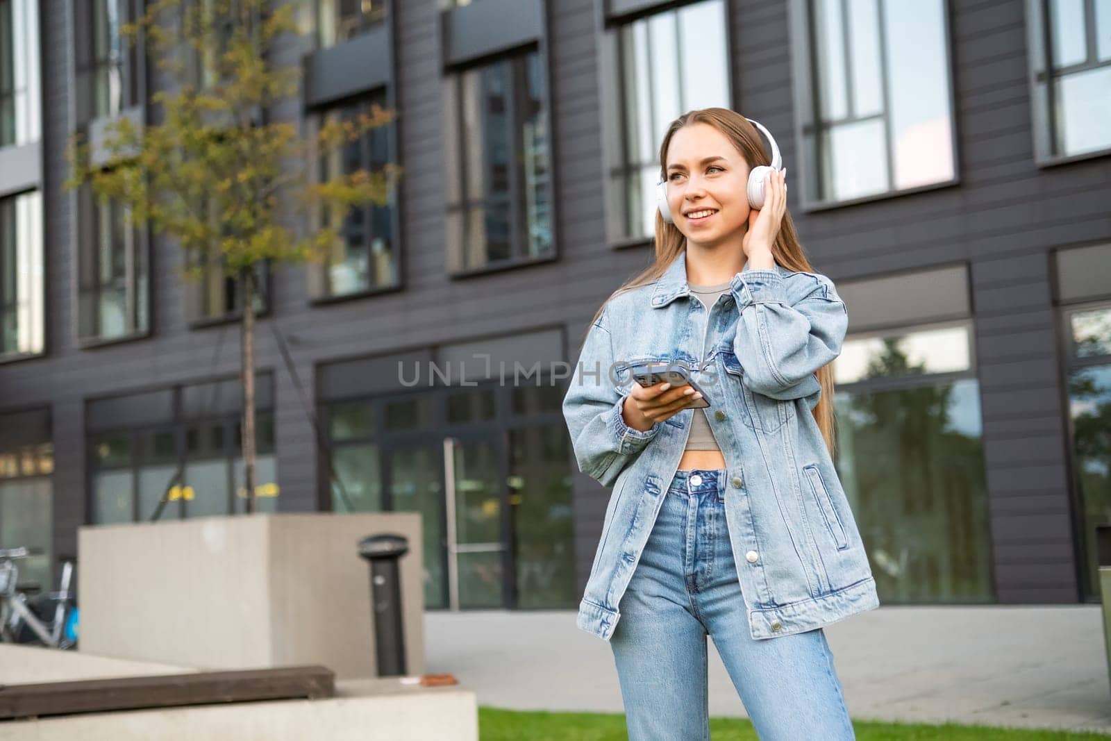 A cheerful young lady, sporting headphones and clutching a mobile device, beams with delight in the street