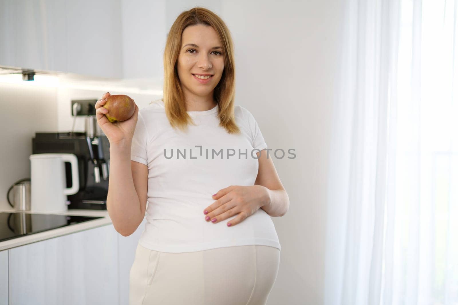 A smiling blonde hair pregnant woman is holding an apple and putting her hand on her belly in the kitchen. Diet during pregnancy.