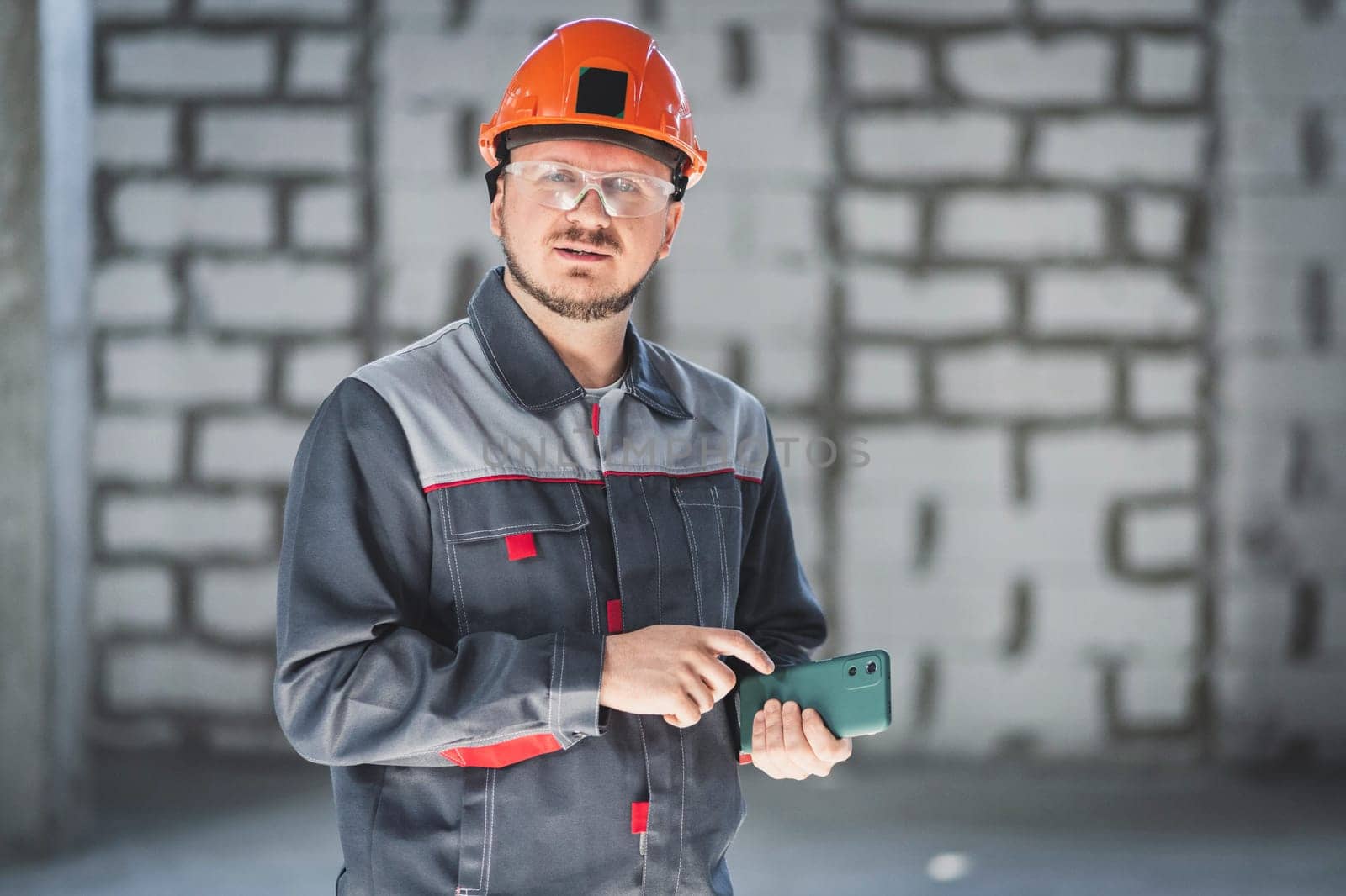 Caucasian construction worker in overalls and helmet uses a smartphone with cameras in his professional work.