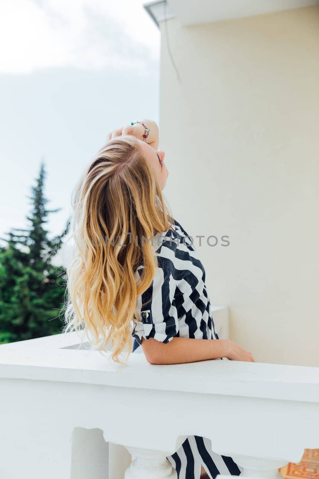 Beautiful fashionable blonde woman in light summer striped dress standing on balcony looking at street