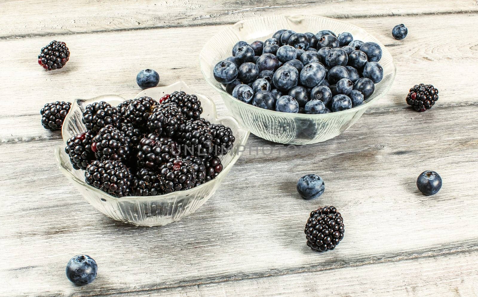 Blueberries and blackberries in glass bowls, some of them spilled on gray wood desk. by Ivanko