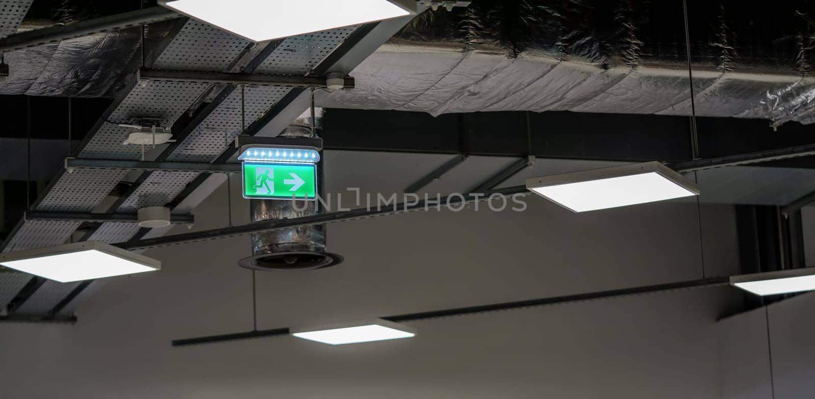 Illuminated green emergency / fire exit sign with running man pictogram on industrial roof. by Ivanko