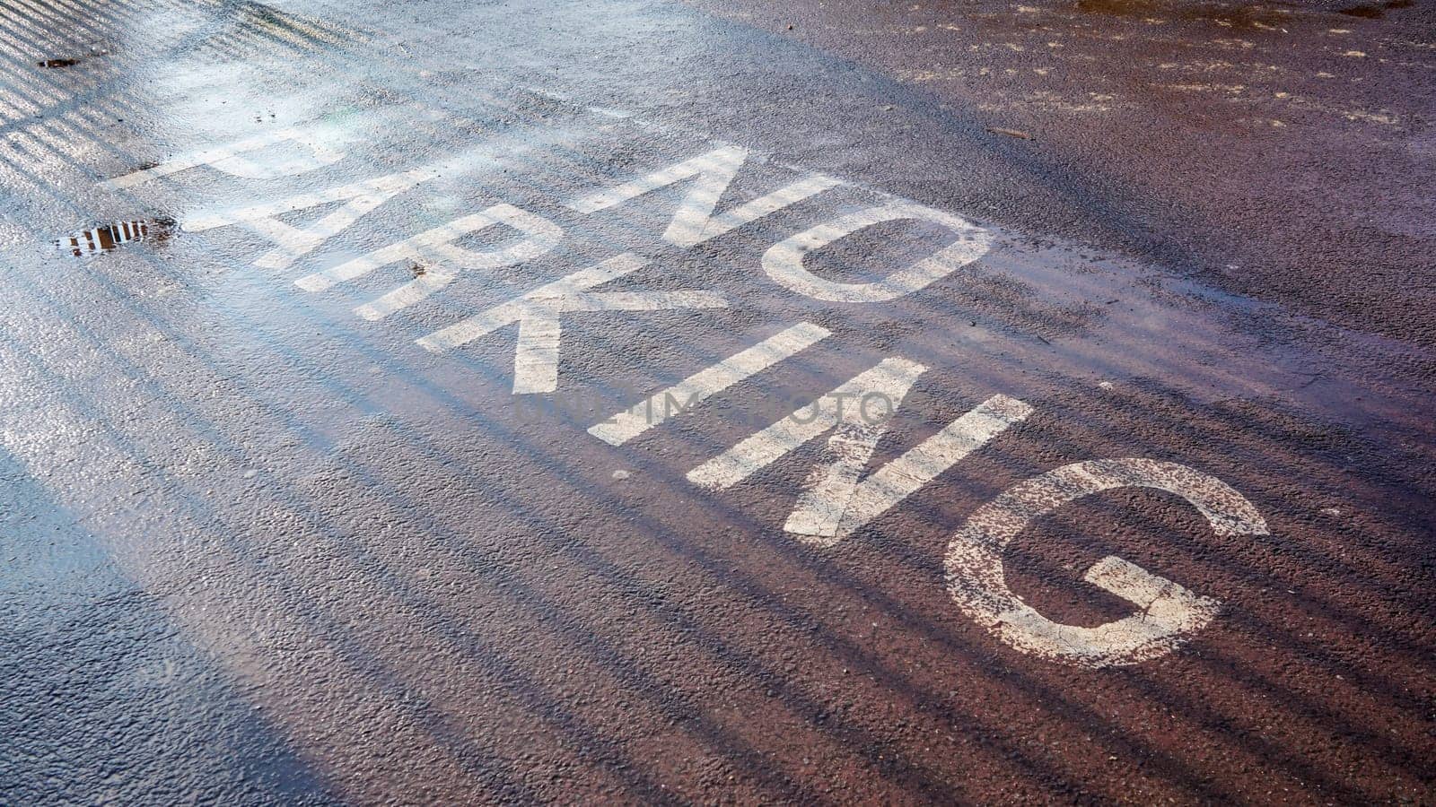 Sun shines on NO PARKING writing on wet asphalt, near fence casting shadow lines by Ivanko