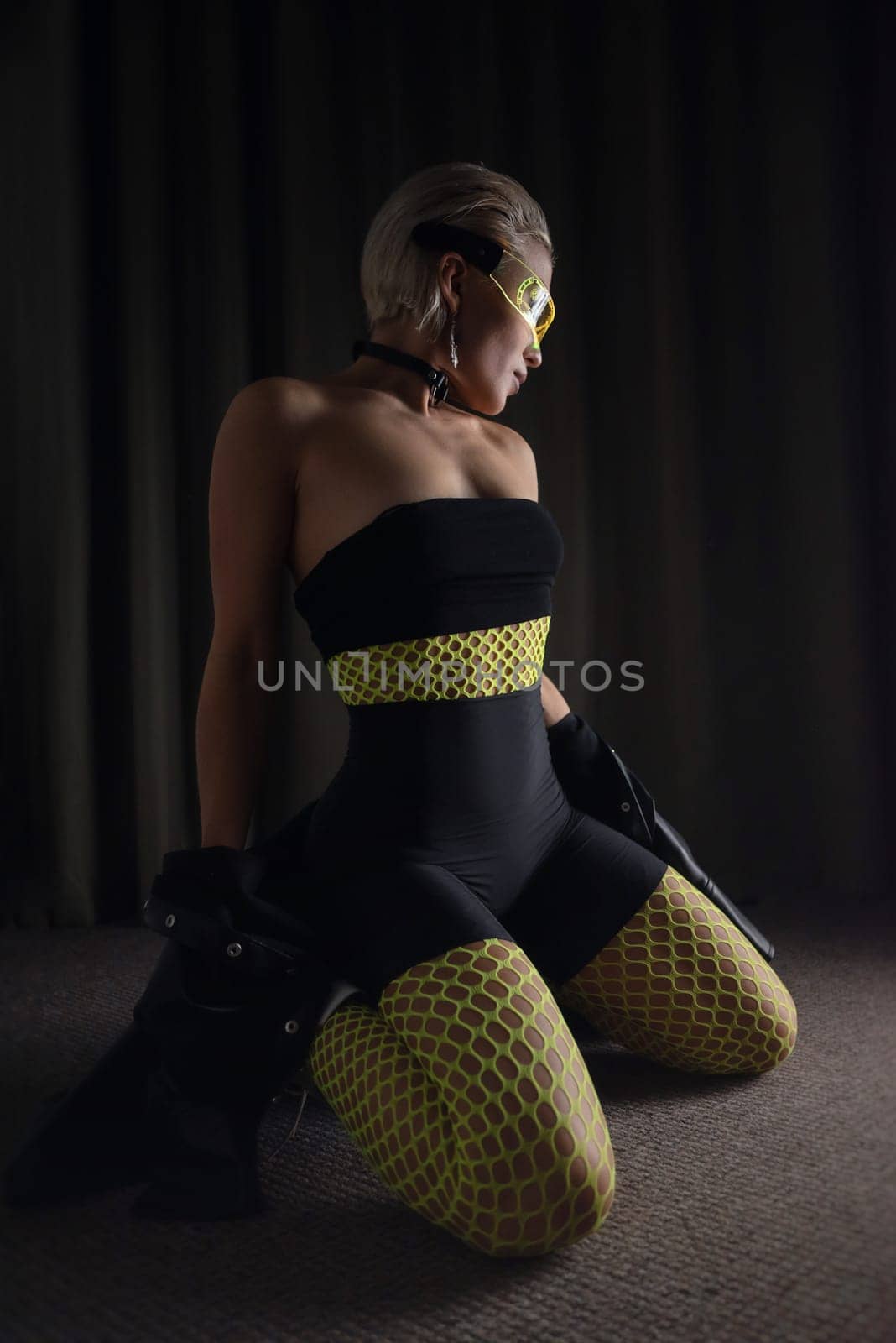 sexy girl in futuristic neon glasses and tight clothes and bright fishnet tights on a dark background in the room