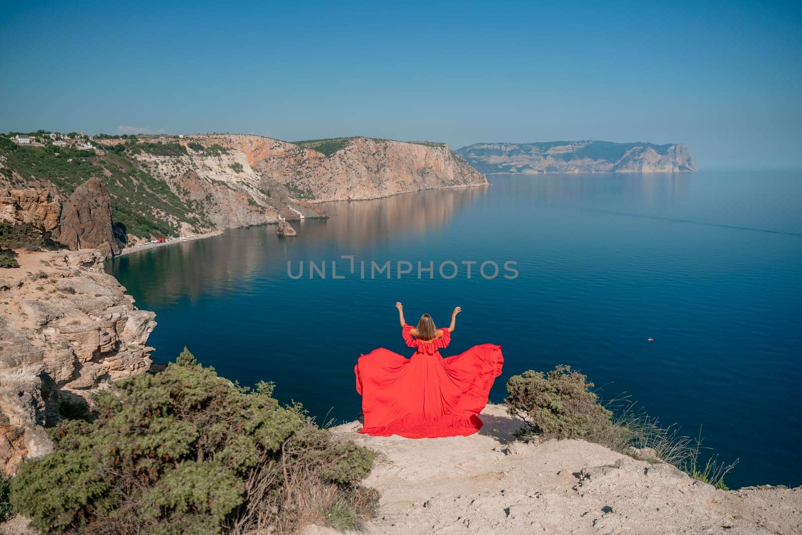 Red dress sea woman. Happy woman with flowing hair in a long flowing red dress stands on a rock near the sea. Travel concept, photo session at sea by Matiunina