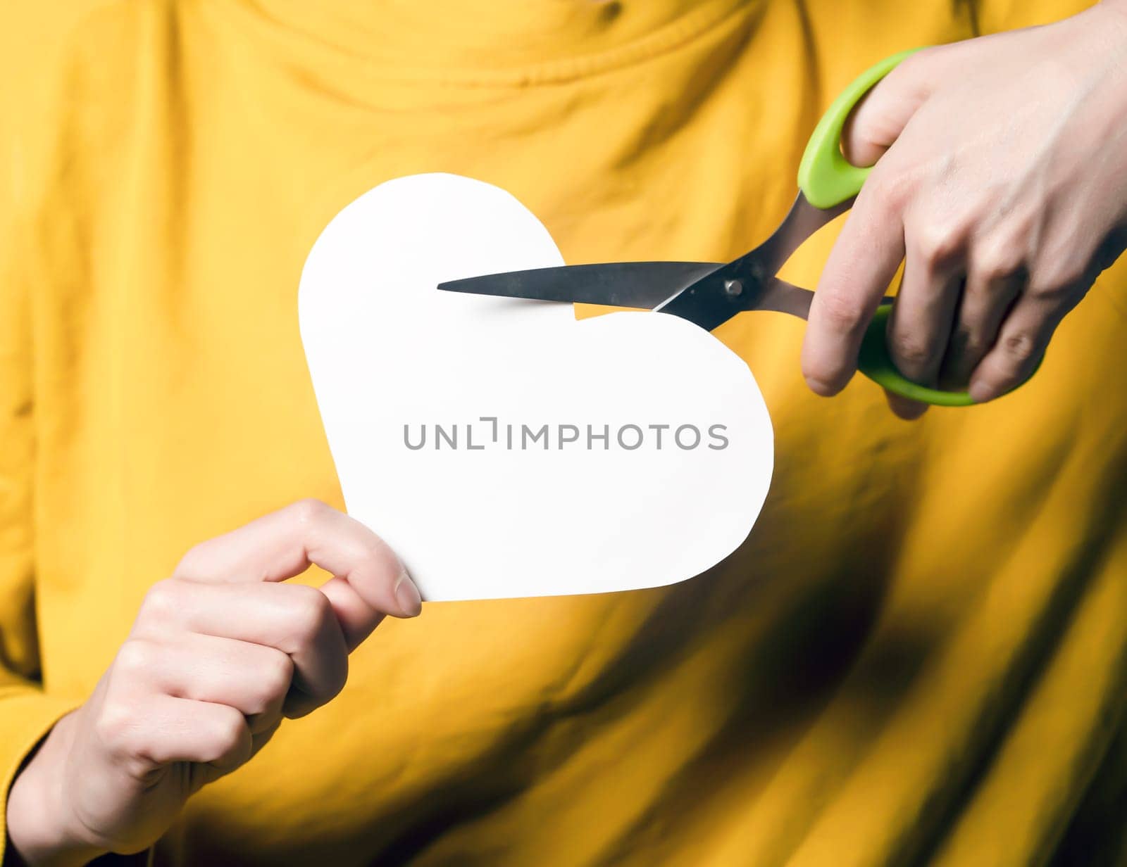 A girl holds scissors in her hands and cuts a heart, a young woman in a yellow sweater expresses her feelings, ends an abusive relationship with toxic love, dissolves a marriage, hands closeup.