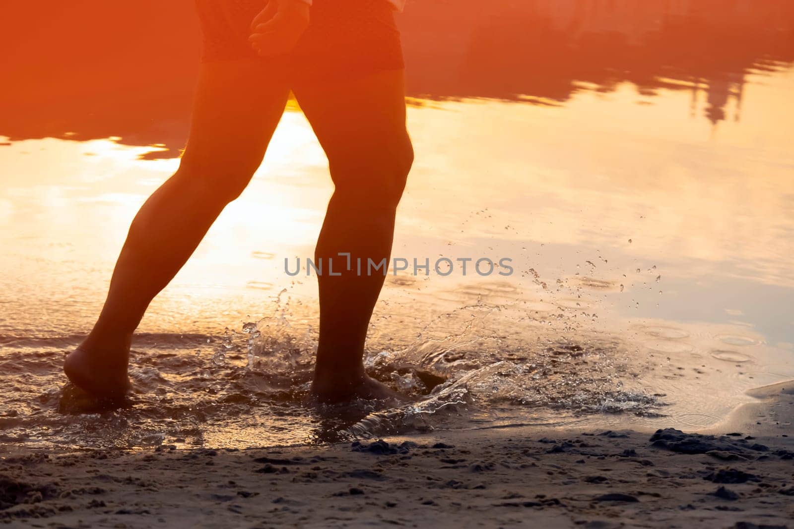 A person runs along the beach on the water, the sea or ocean in the rays of the sun, an athlete man spends time happily, is training, jogging outdoors, close-up view of the legs and splashes.