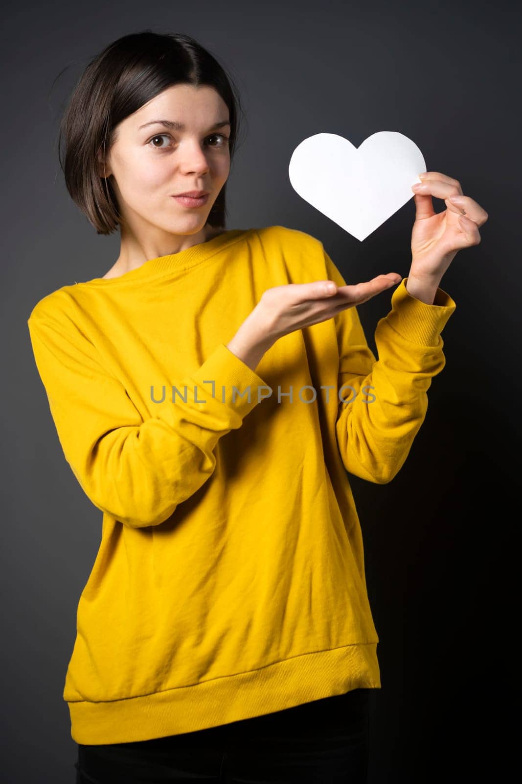 A young girl holds a white paper heart - a postcard or shows feelings of love. A woman with a joyful surprised face before the holiday and in search of love, relationships or shares her impressions.