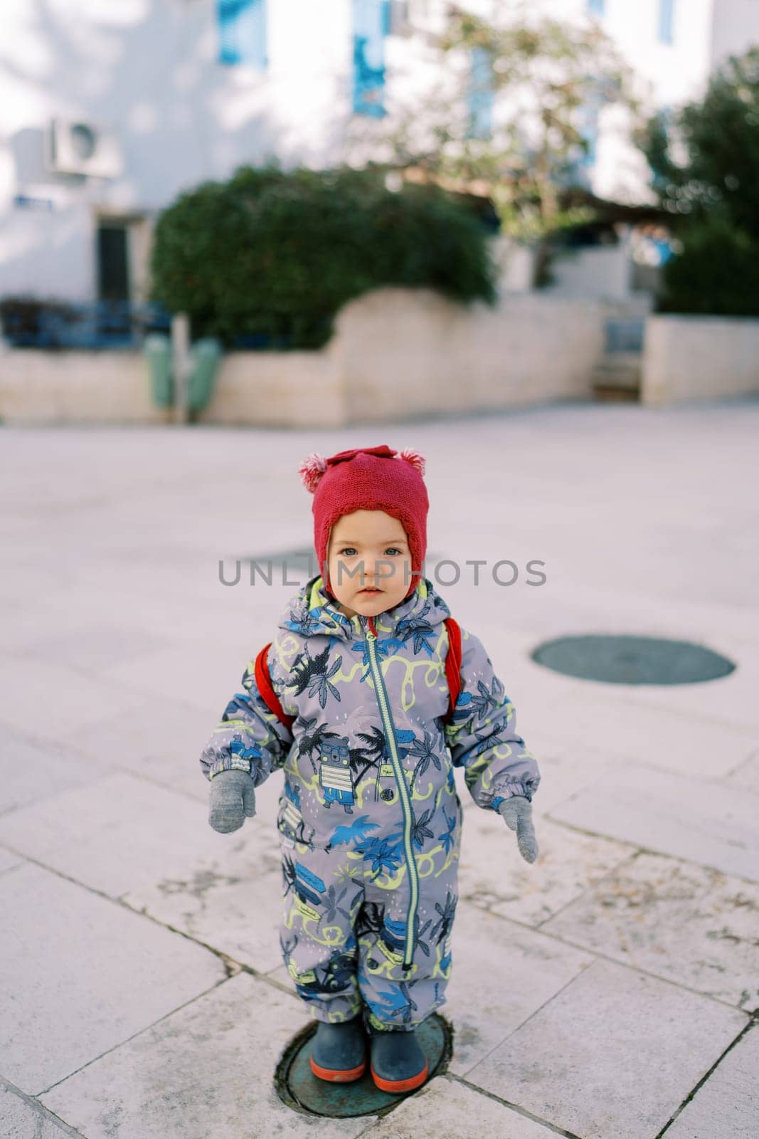 Little girl in overalls and a hat stands on a tile in the courtyard of the house. High quality photo