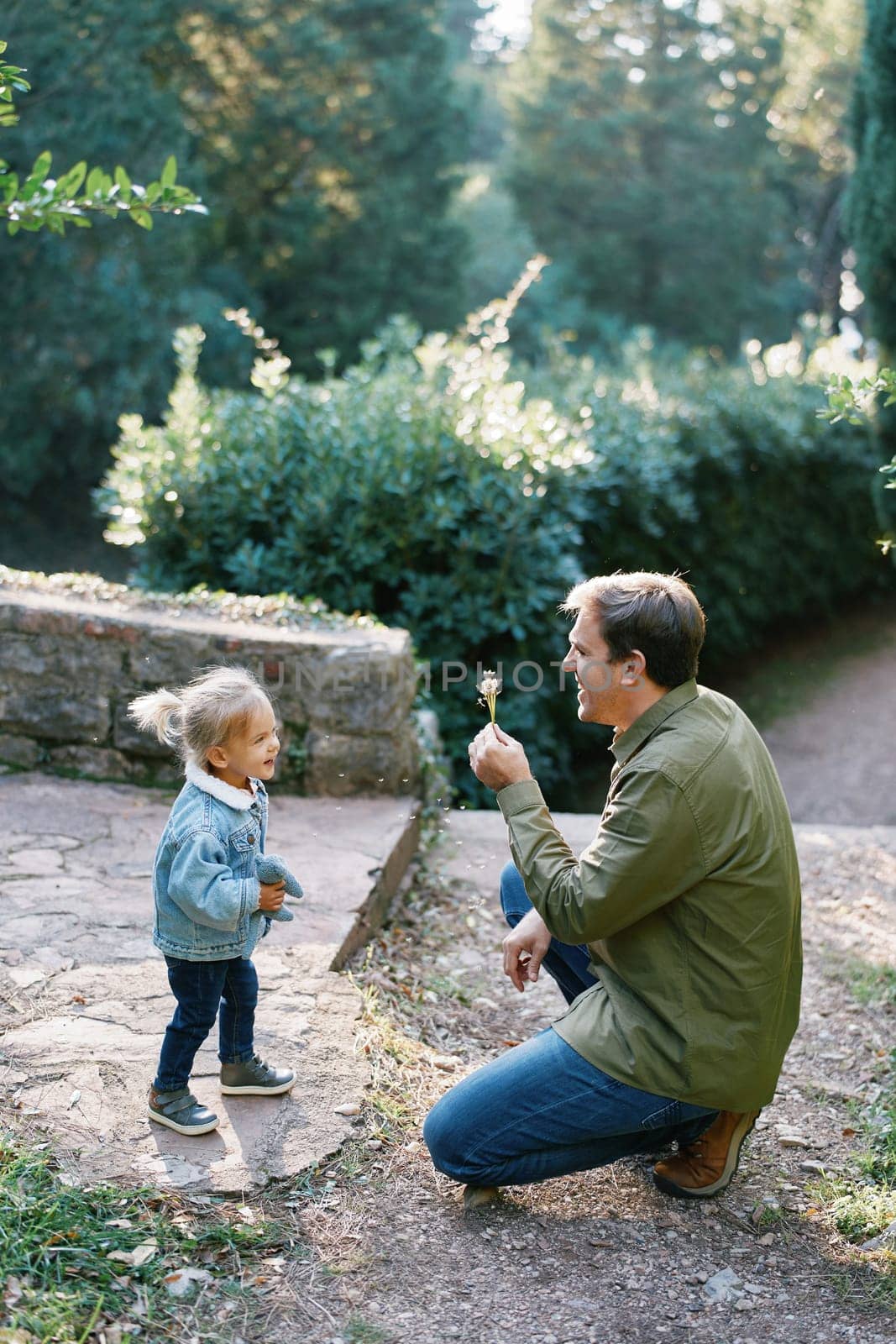 Little smiling girl stands in the park near a crouched dad with a bouquet of dandelions. High quality photo