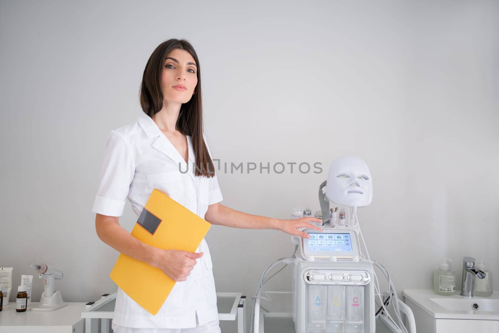 Cosmetologist standing in medical office. Female dermatologist holding yellow folder standing and look in camera near modern equipment. Dermatology specialist in uniform at hospital