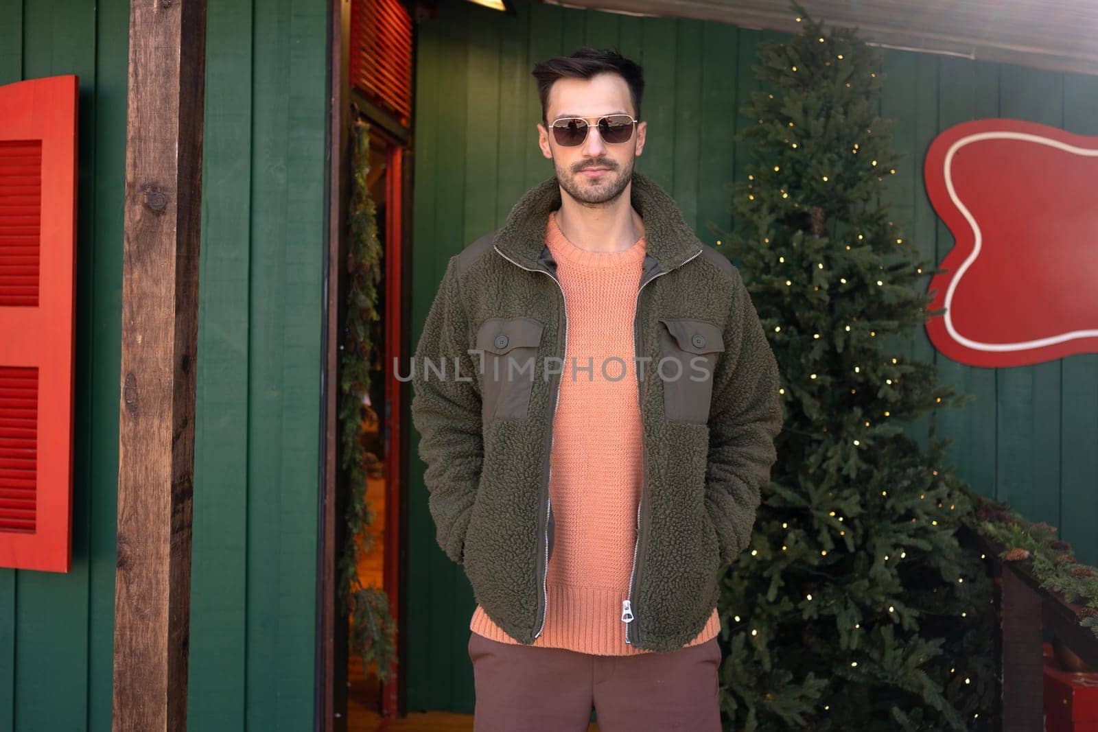 Man wear sunglasses standing near Christmas tree. Young male with trendy hairstyle wearing green jacket and pink shirt stands on street near storefront with bright lights