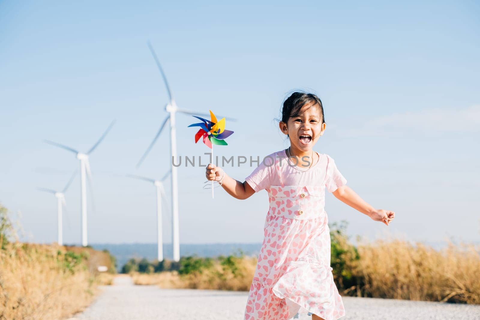 Child's joyful play near windmills, little girl runs with pinwheels. Embracing playful wind energy education and clean electricity in a picturesque sunny wind turbine setting.