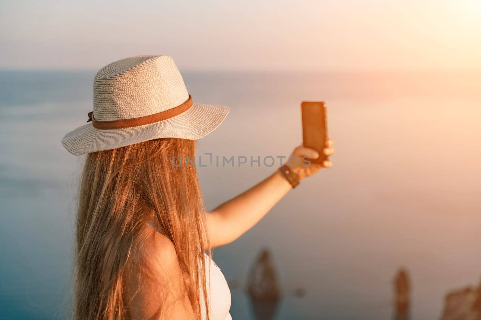 Selfie woman in a hat, white tank top, and shorts captures a selfie shot with her mobile phone against the backdrop of a serene beach and blue sea