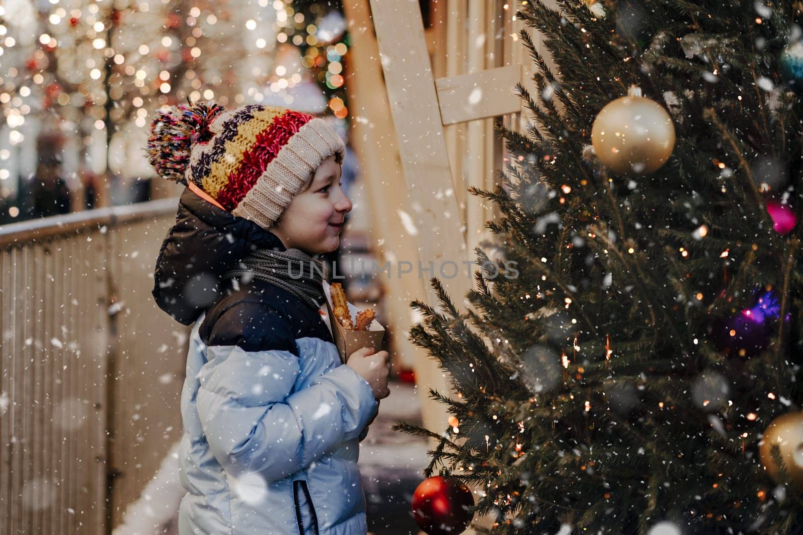 Cute little boy eating churros on traditional Christmas fair in snow. Child kid enjoying sweets on Xmas market. Winter holidays with family. Merry Christmas.