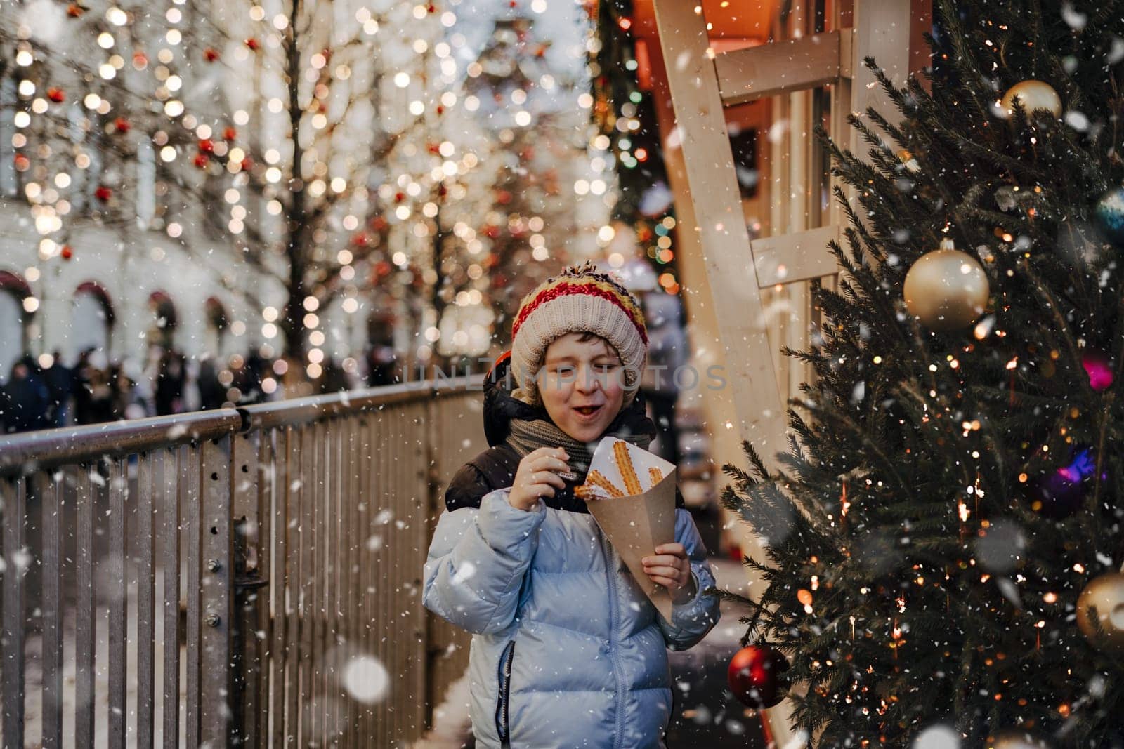 Cute funny little boy eating churros on traditional Christmas fair in snow. Child kid enjoying sweets on Xmas market. Winter holidays with family. Merry Christmas by Ostanina