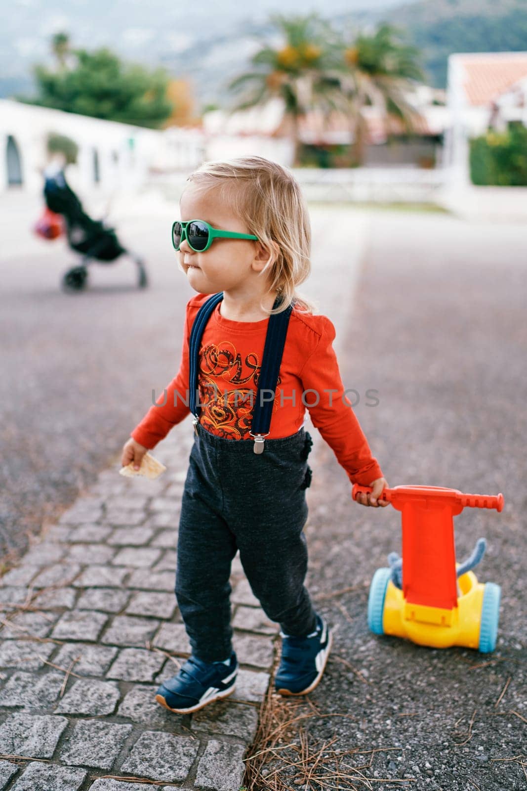 Little girl in sunglasses drags a toy cart along the cobblestones. High quality photo