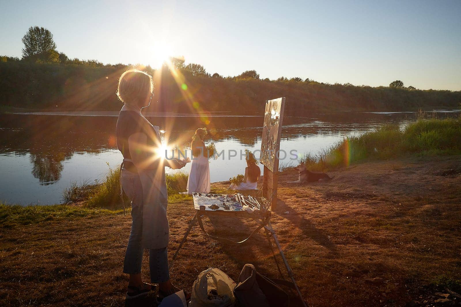 Adult female artist painting picture near water of river or lake in nature and girls in white sundress and flower wreath posing in the holiday of Ivan Kupala in nature at sunset by keleny