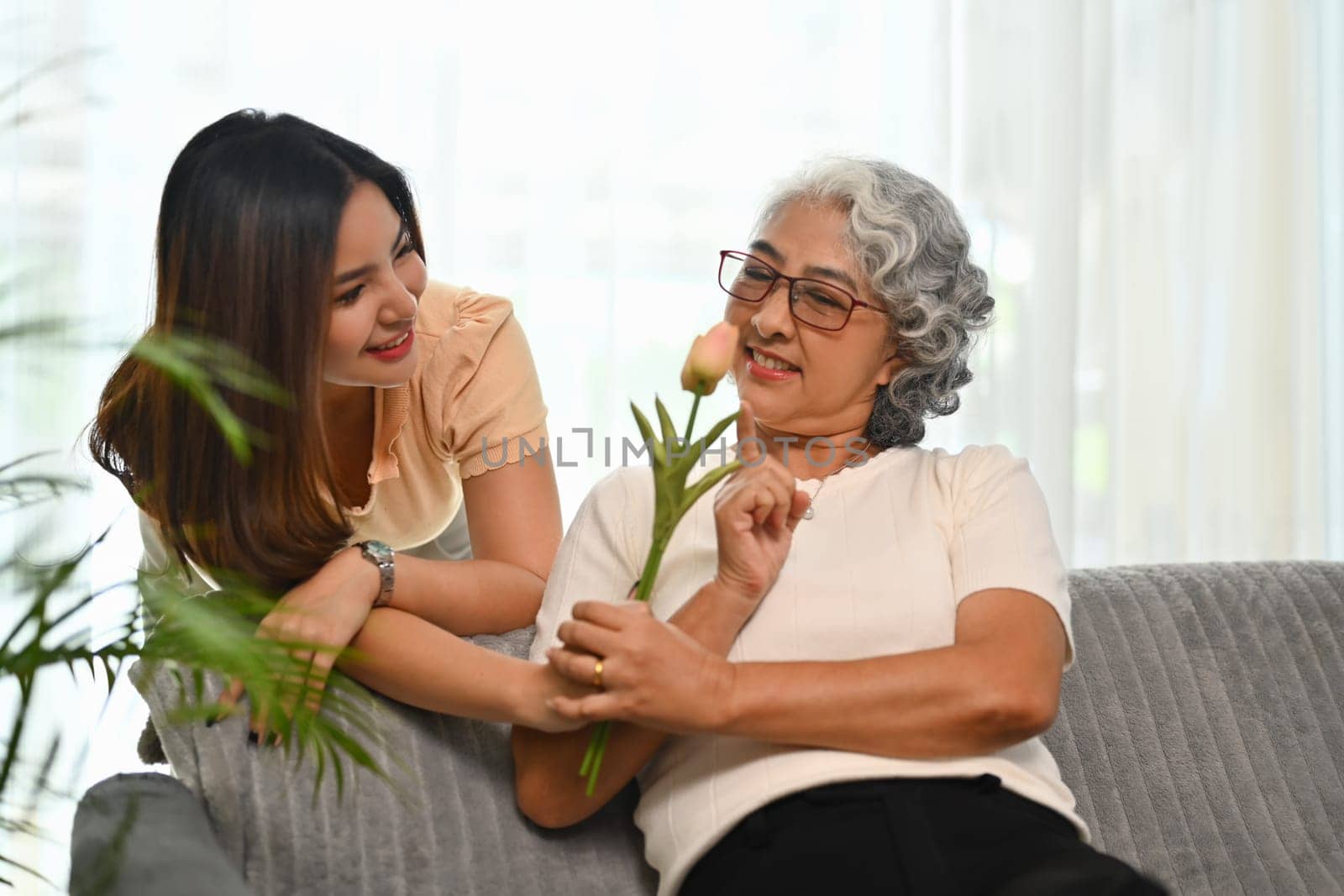 Adult daughter standing behind couch and giving flowers to older mother. Family bonding concept.