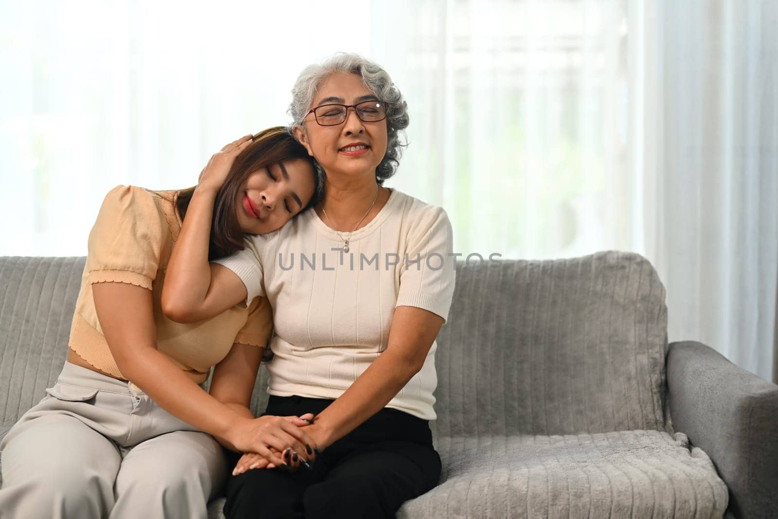 Beautiful grey haired elderly woman embracing her grownup daughter sitting on sofa at home.