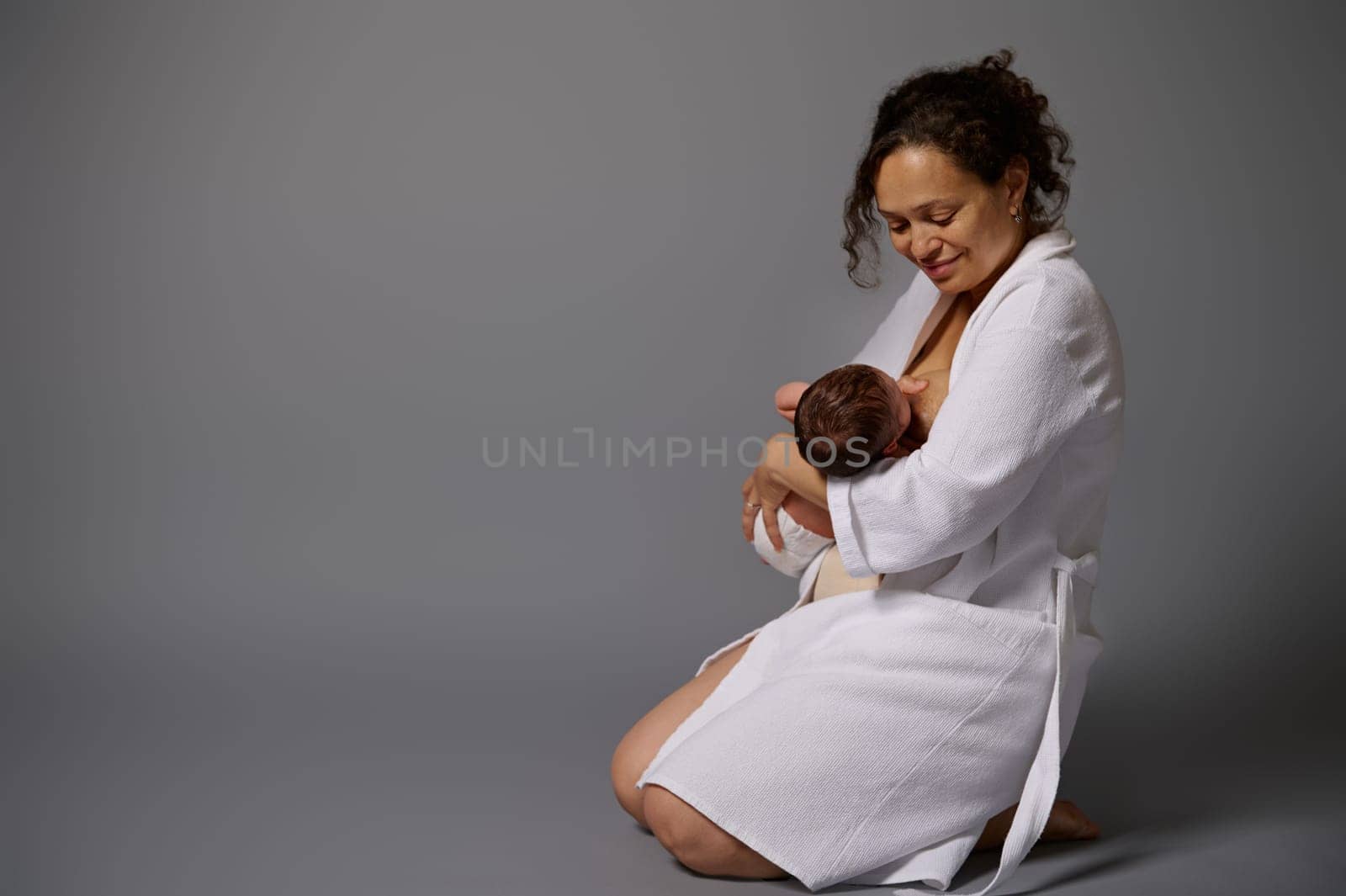 Full length portrait of a peaceful loving young African American mother in bathrobe, feeding her newborn baby with breast, isolated on gray studio background with copy ad space. Mom nursing baby.