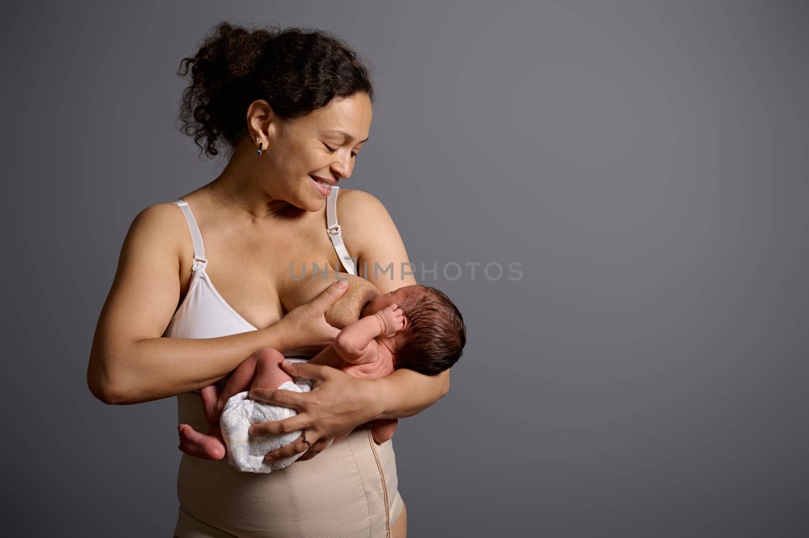 New mother feeds her newborn baby with breast milk, for healthy growth, smiling isolate on gray studio background with copy advertising space, dressed in underwear and elastic bandage after c-section
