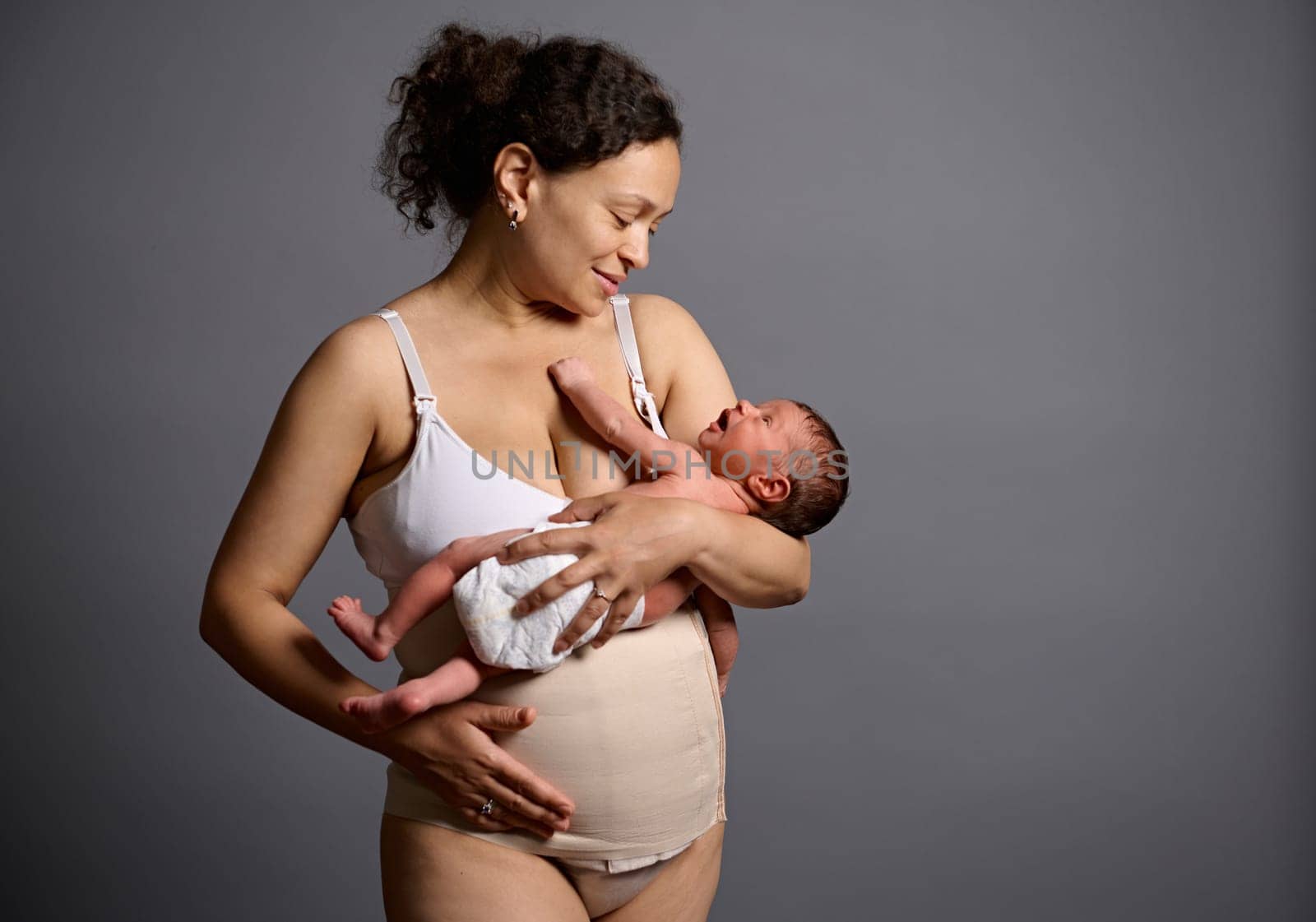African happy mother in underwear and elastic bandage on her belly after c-section, holding and smiling looking at her newborn baby, isolated over gray studio background with free advertising space