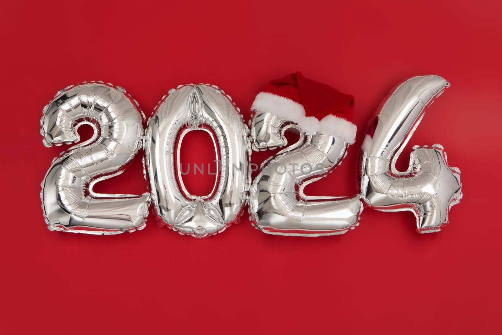 2024 inflatable balloons on red background with Santa hat. Happy New year twenty twenty four celebration. Silver foil balloons numeral 2024. Postcards and posters Copy space