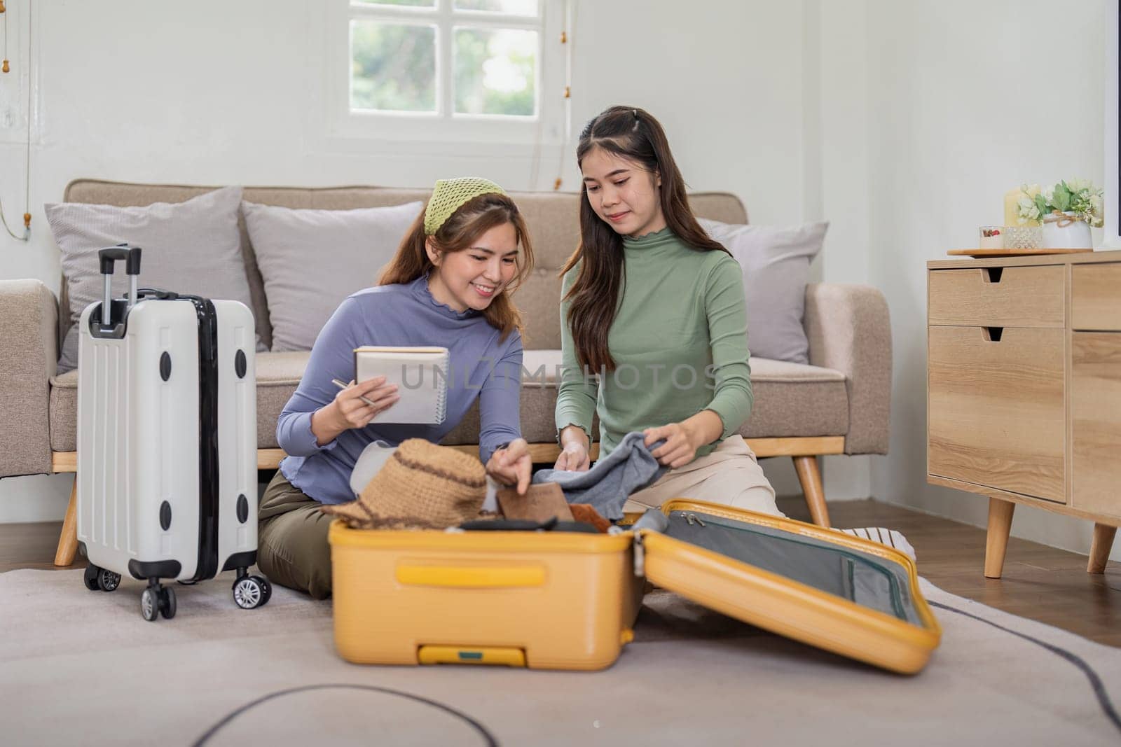 Two young Asian women are prepare for the journey happily. Pretty girls are packing suitcase for the trip and check the list of various items.