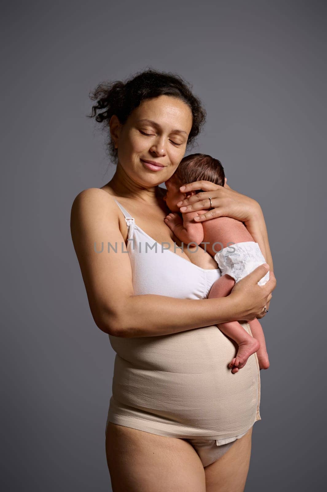 Multiethnic young woman, a loving caring mother, dressed in underwear and elastic bandage after c-section cesarean, carrying and hugging her newborn baby, isolated on gray studio background. Ad space