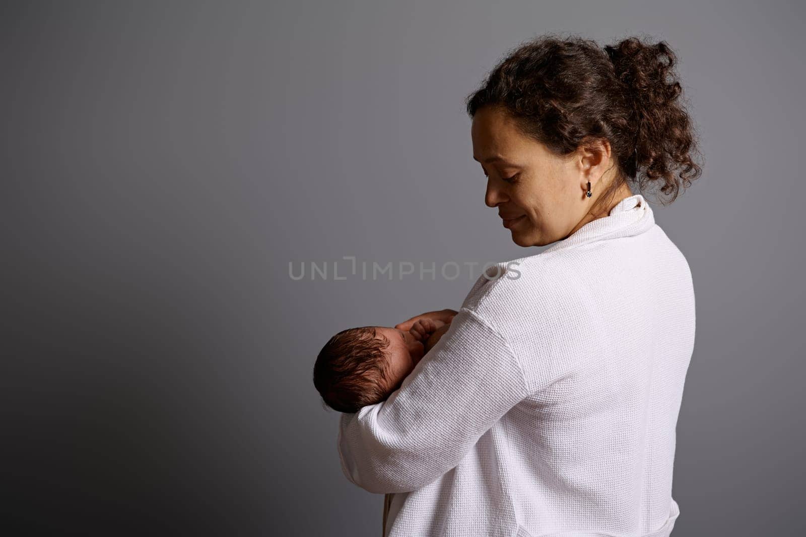 Rear view of a happy woman, young mother, breastfeeding her newborn baby, isolated on a gray studio background. Milk from mother's breast is a natural medicine for children. Copying advertising space