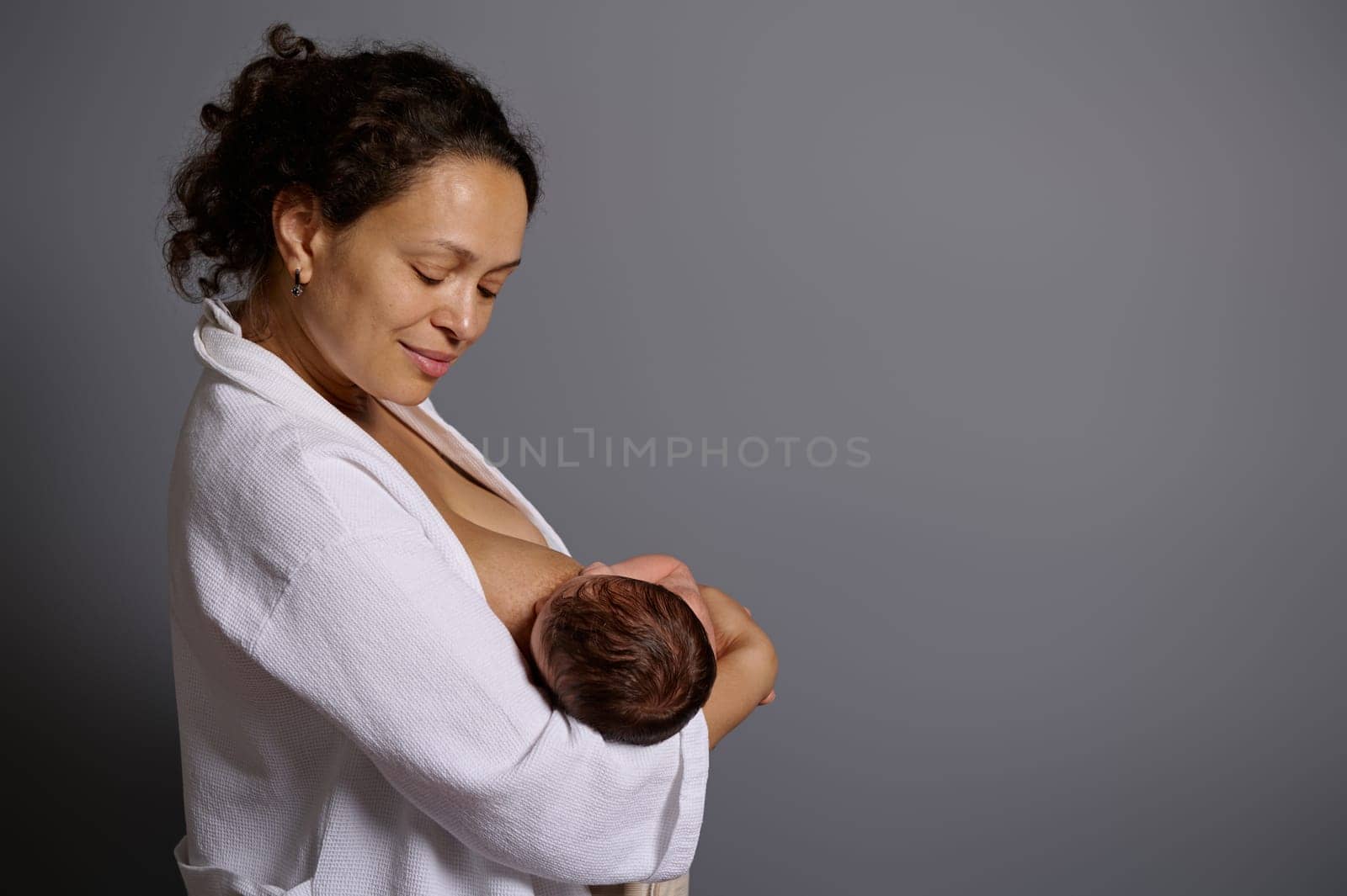 Smiling beautiful woman enjoys breastfeeding her baby, isolated over gray studio background. The importance of breast milk for newborns for first six month of life. Maternity leave and babyhood period