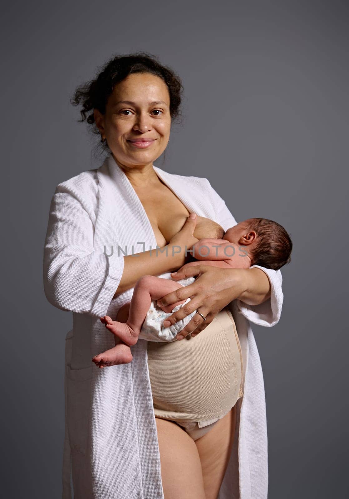 Authentic curly haired multi ethnic woman, happy young mother looking at camera, holding her newborn baby suckling at her breast, feeding him with healthy wholesome breast milk, isolated on gray