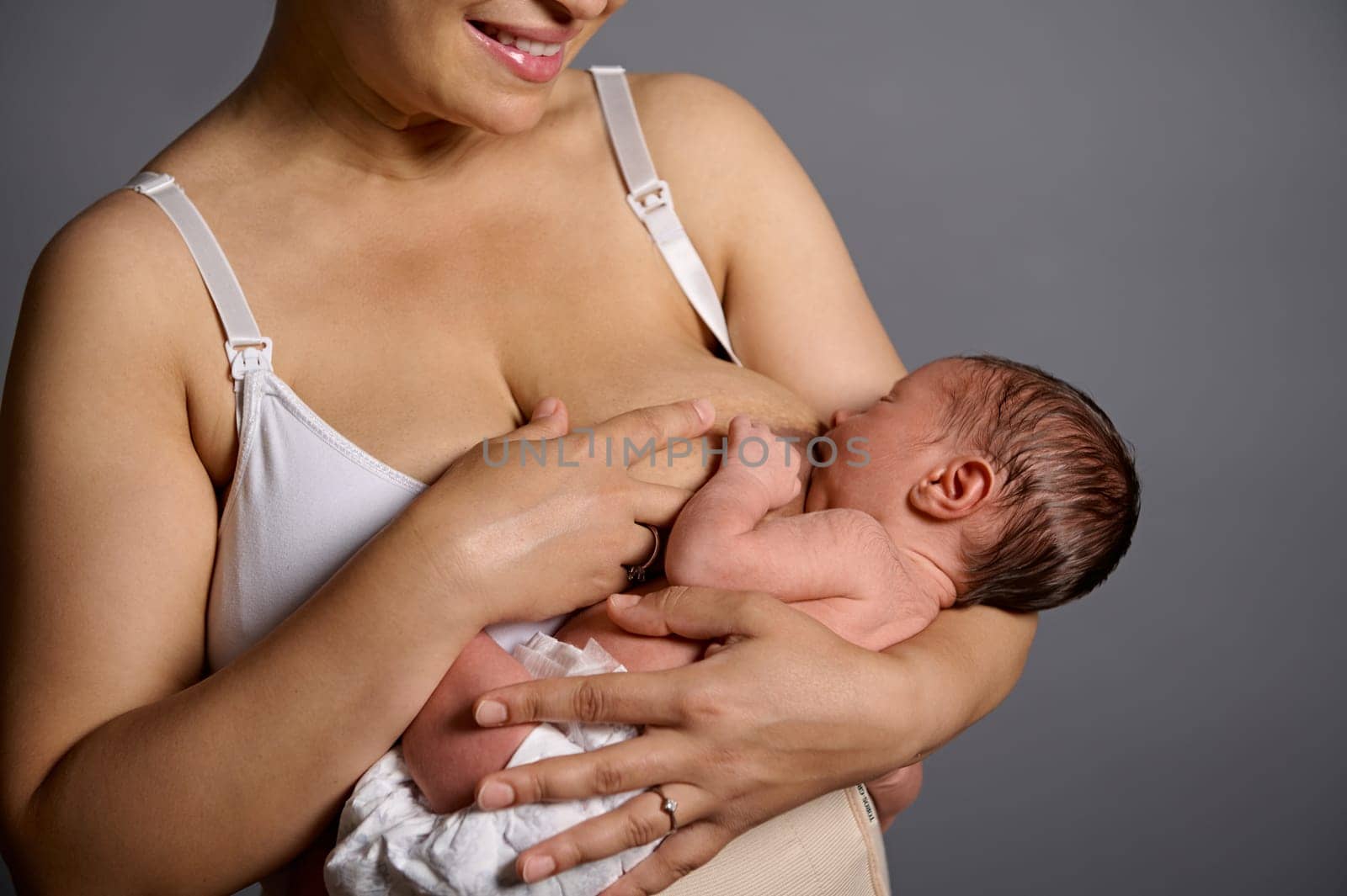 Cropped view of young smiling new mother feeding her newborn baby with breast milk, for healthy growth, gray studio background. Copy ad space, dressed in underwear and elastic bandage after c-section