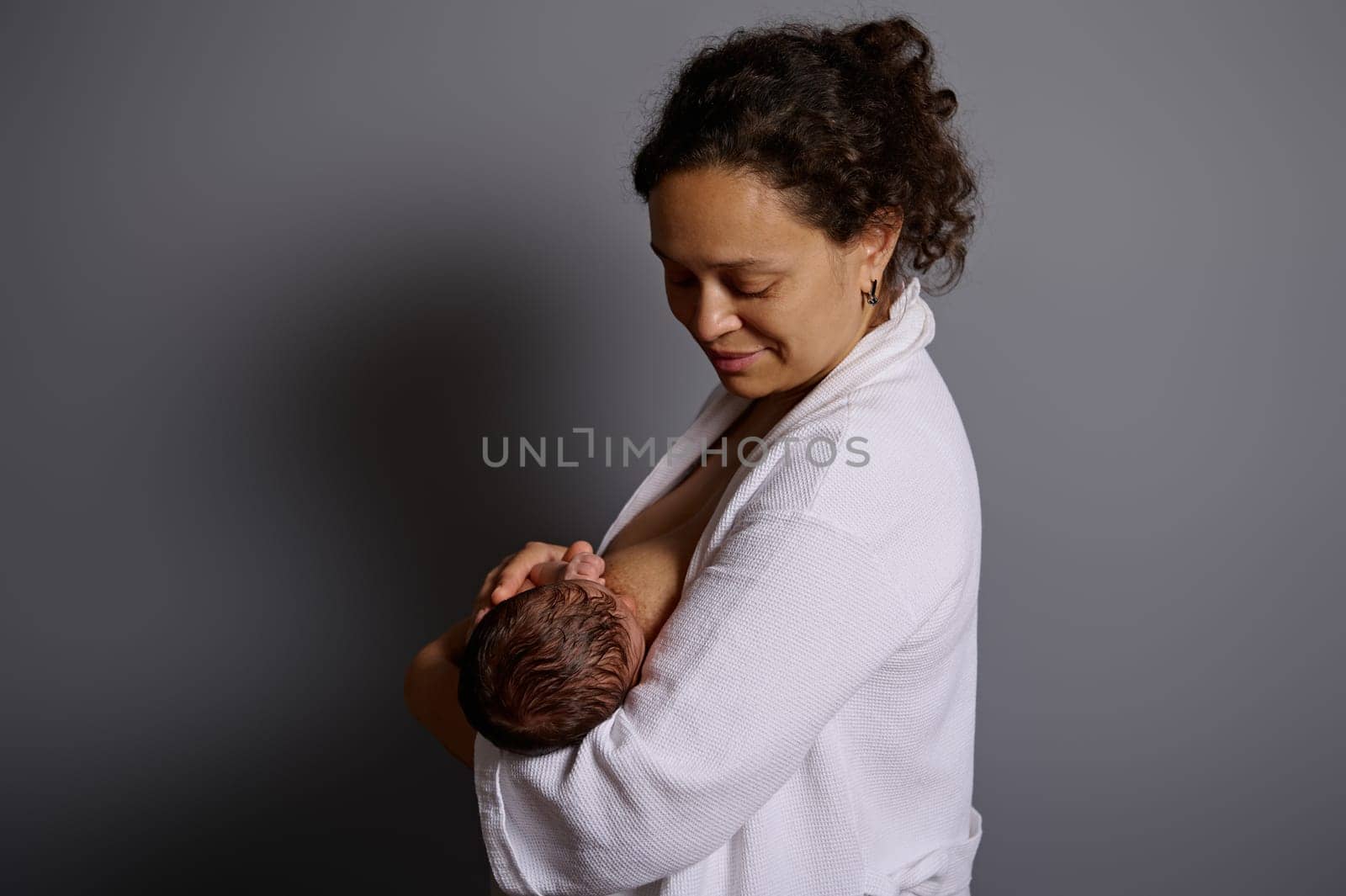 Authentic studio portrait of African American pretty woman, young mother smiling looking at her newborn baby, while breastfeeding him. People. Maternity leave. Healthy lifestyle