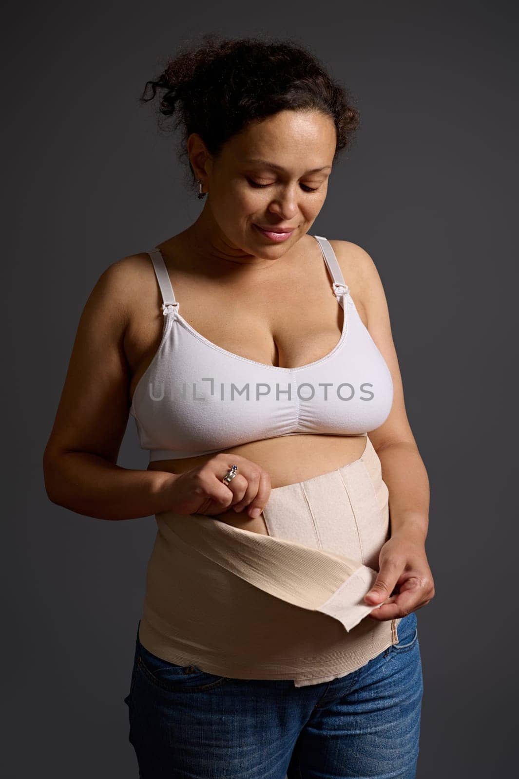 Middle aged woman post pregnancy and childbirth, putting on an elastic bandage after surgical operation cesarean section, isolated over gray background. Postpartum recovery period. Skin and body care